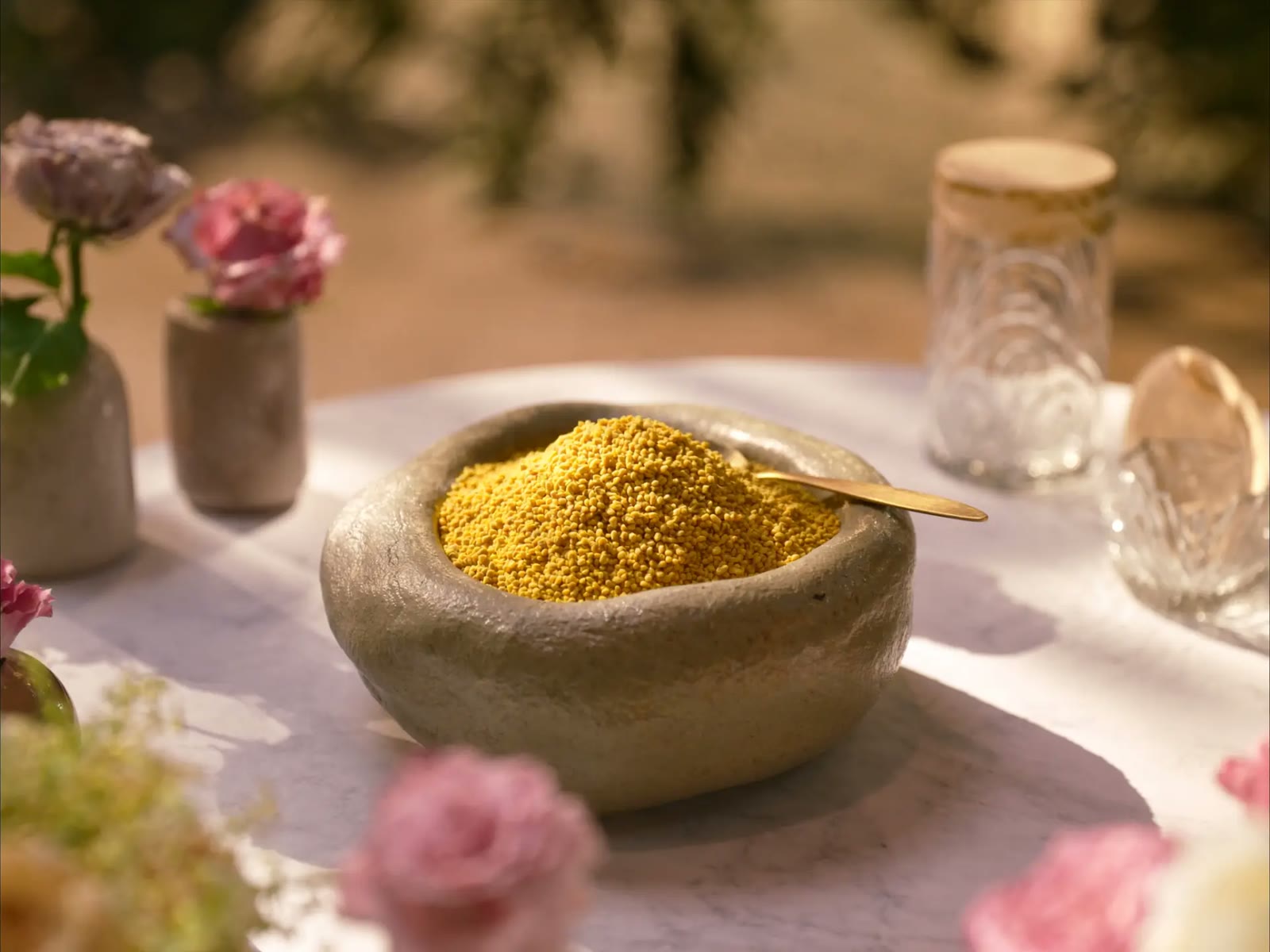 Stone bowl of golden bee pollen with a brass spoon on a sunlit table with pink roses
